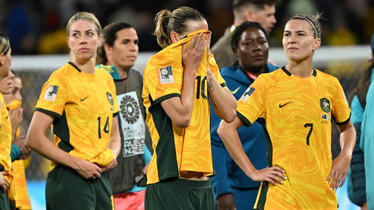 Three Matildas players standing on a stadium field. One is rubbing her face with her top.
