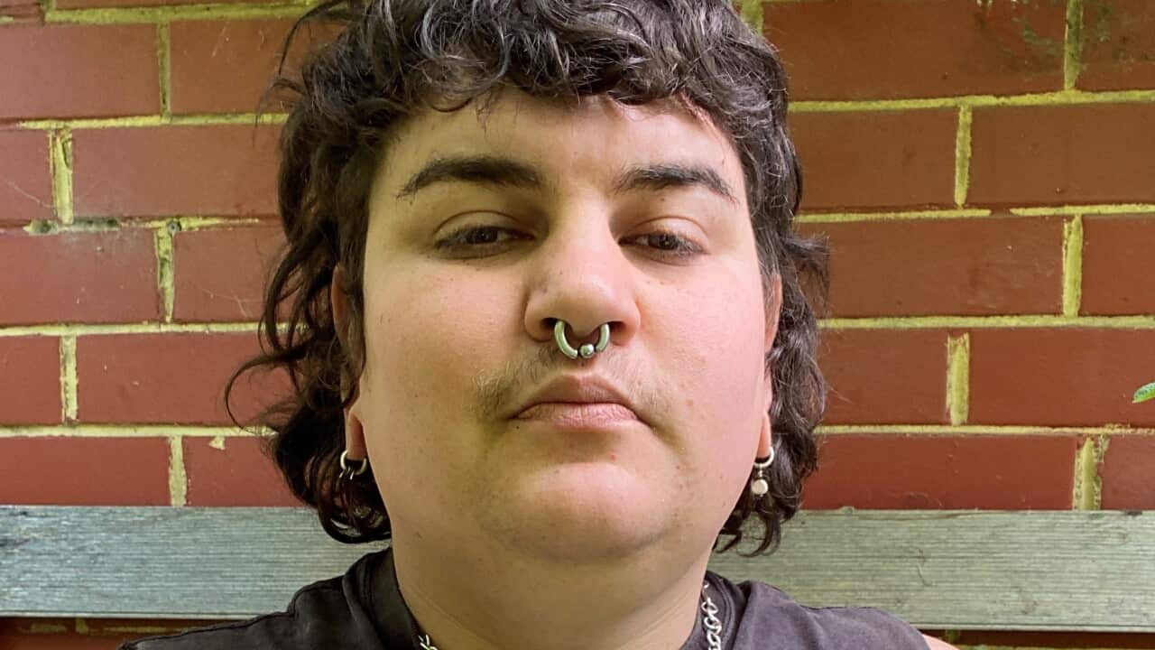 A person with short dark hair, wearing a black tank top, standing in front of a brick wall.