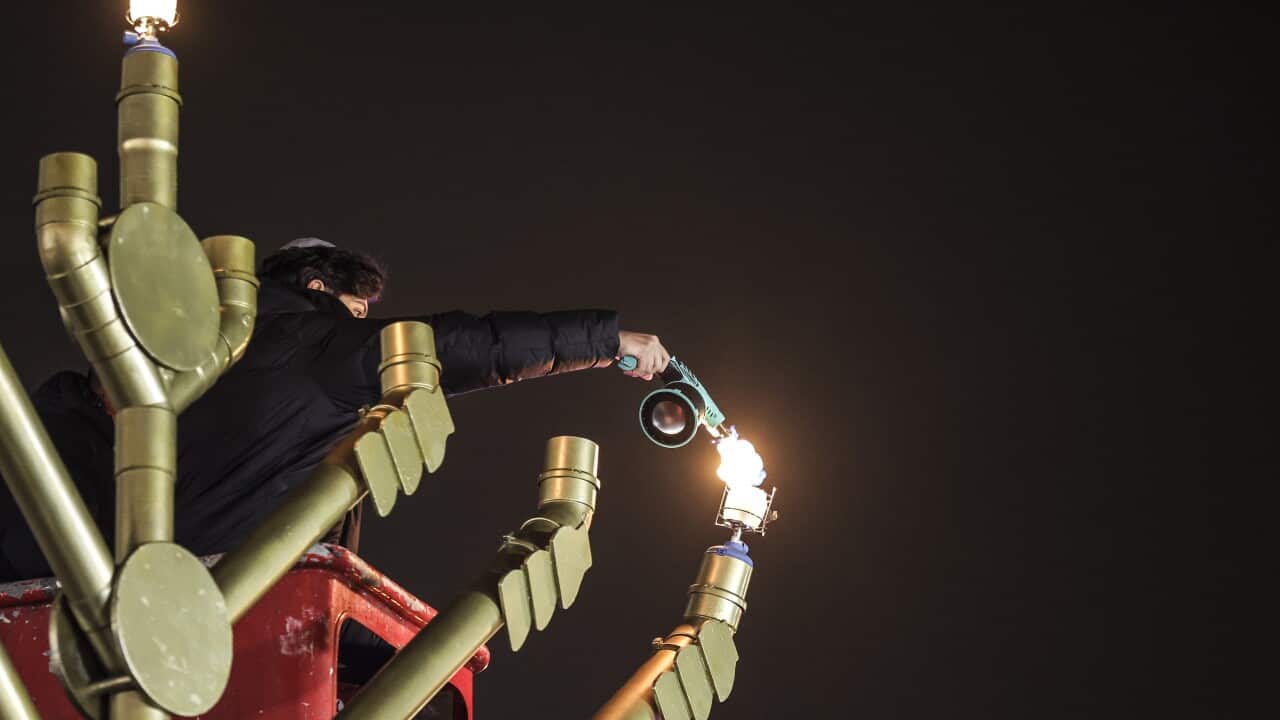 A boy wearing a black jacket and yarmulke lights the arm of a menorah larger than himself.