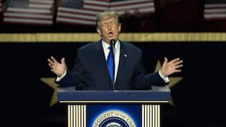 Donald Trump speaks passionately with open hands at a presidential podium in front of a backdrop showing American flags.