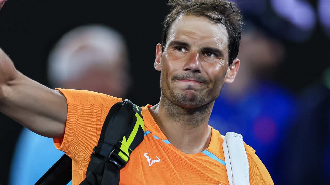A man with bags over his shoulders waves as he walks across a tennis court.