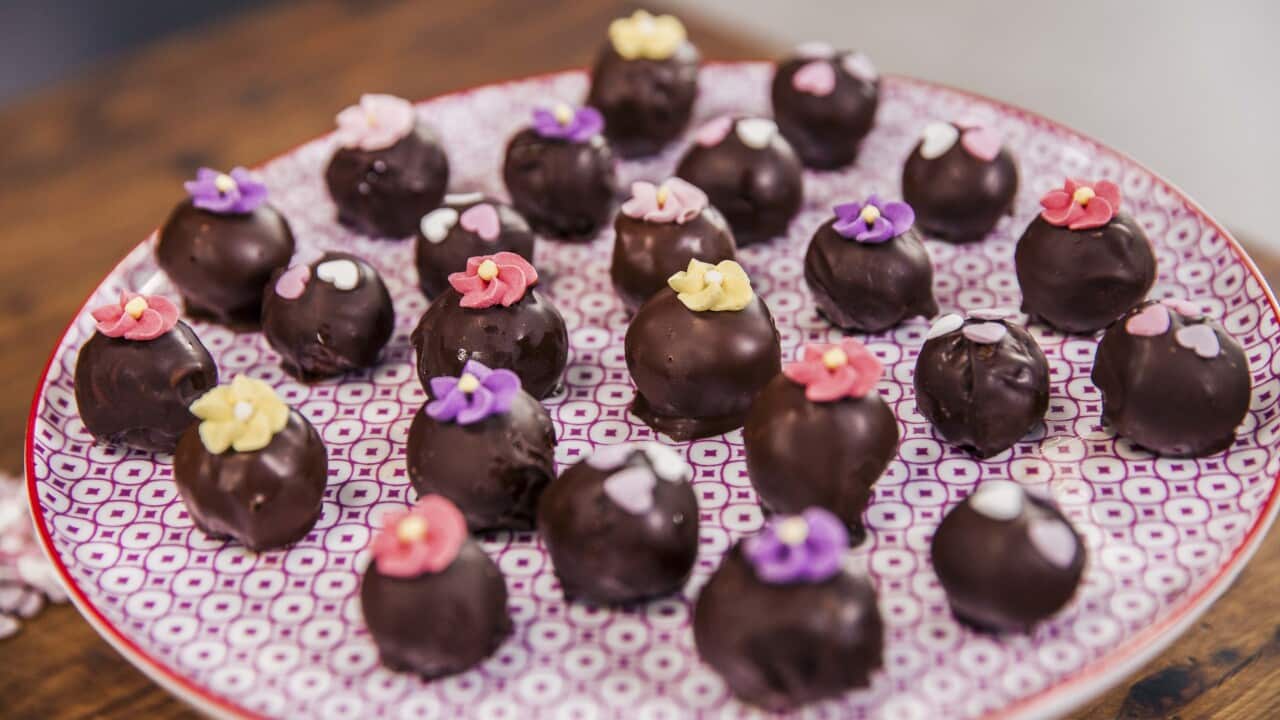 24 chocolate-coated truffles sit on a patterened plate. Some are decorated with small icing flowers.