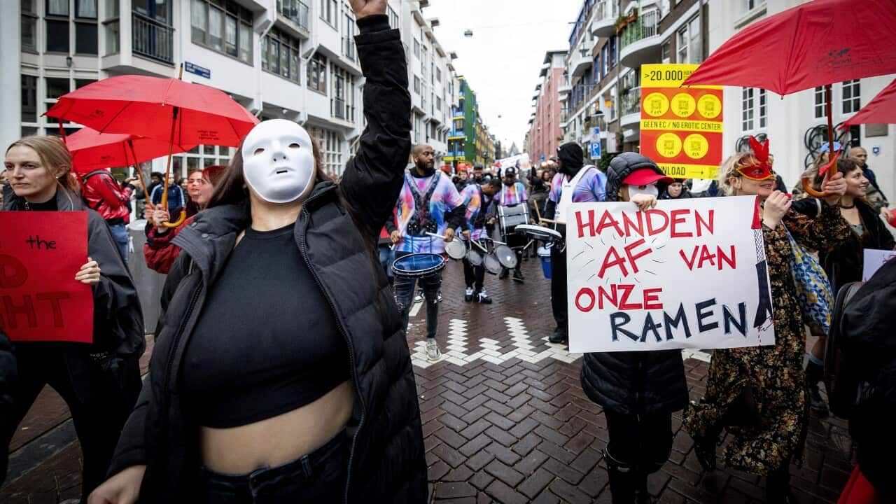 Group of protesters march through streets with red umbrellas, some holding signs. Woman at front of group wears a white mask to conceal her face.