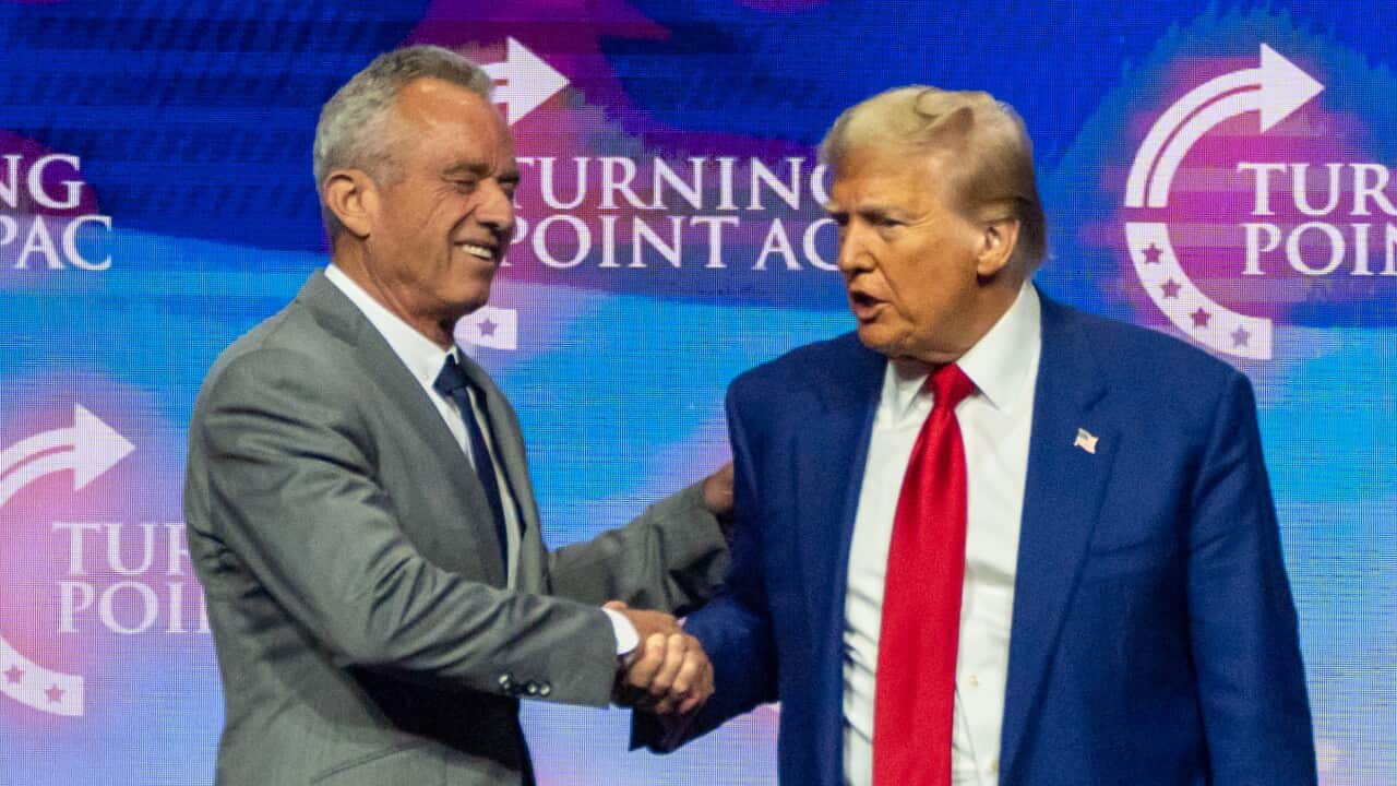 Two men in suits shaking hands on a brightly lit stage with a blue background.