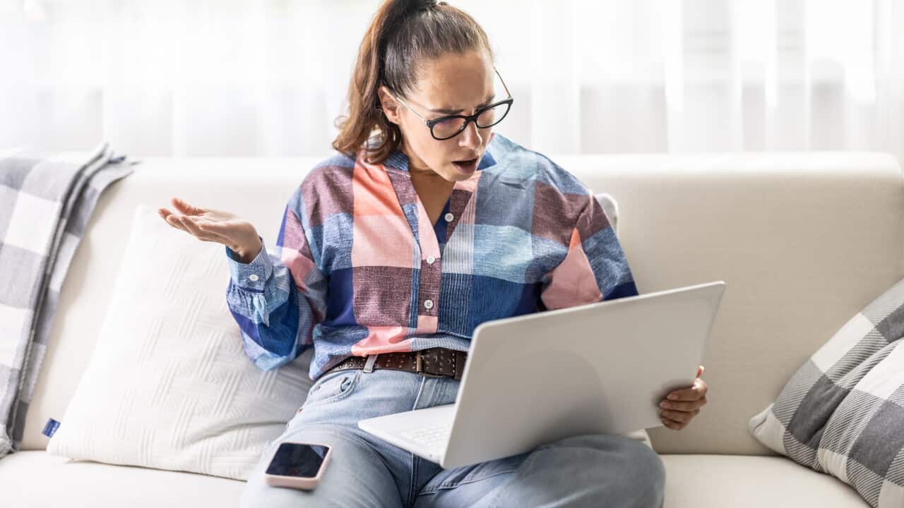 A woman stares at her laptop with her mouth open and a blank expression, throwing her hand up in surprise. Her frustrated reaction and smartphone resting on her thigh suggest that she has just encountered an unexpected problem or bad news.