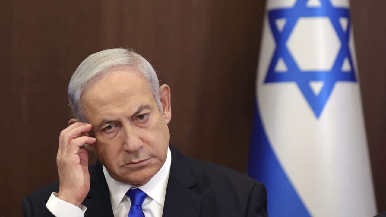 Benjamin Netanyahu, a man with grey hair and wearing a suit, touches his face. The Israeli flag is behind him.