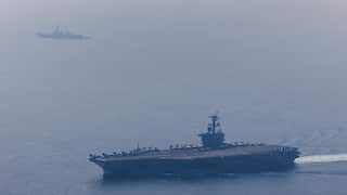 A large aircraft carrier laden with fighter jets sails with a battleship across a calm sea.