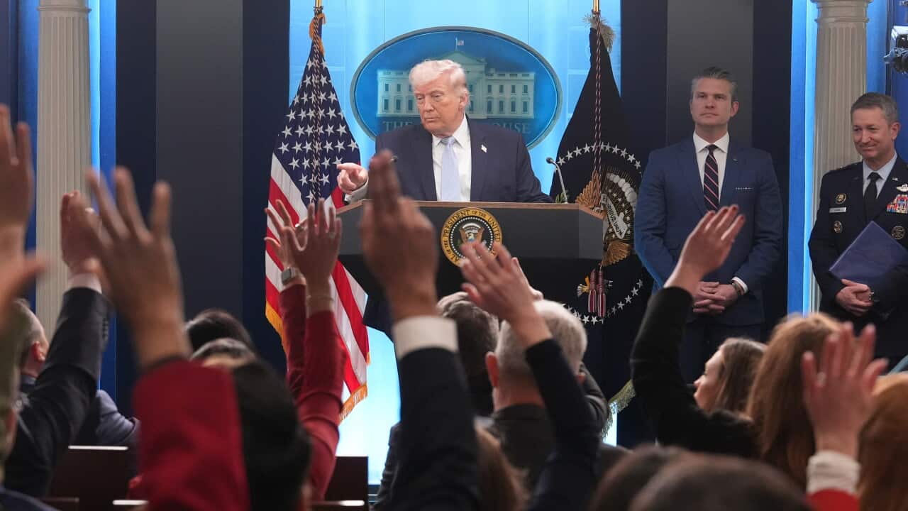 Donald Trump speaks from behind a White House lectern, pointing toward a crowd of journalists with raised hands during a press briefing.