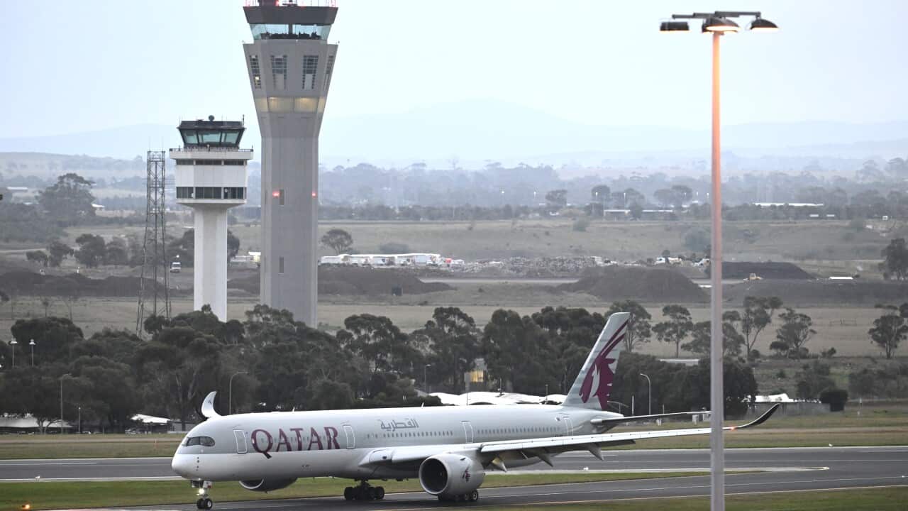 A Qatar Airways plane taxis along a runway in front of air traffic control towers and a grey, overcast background.