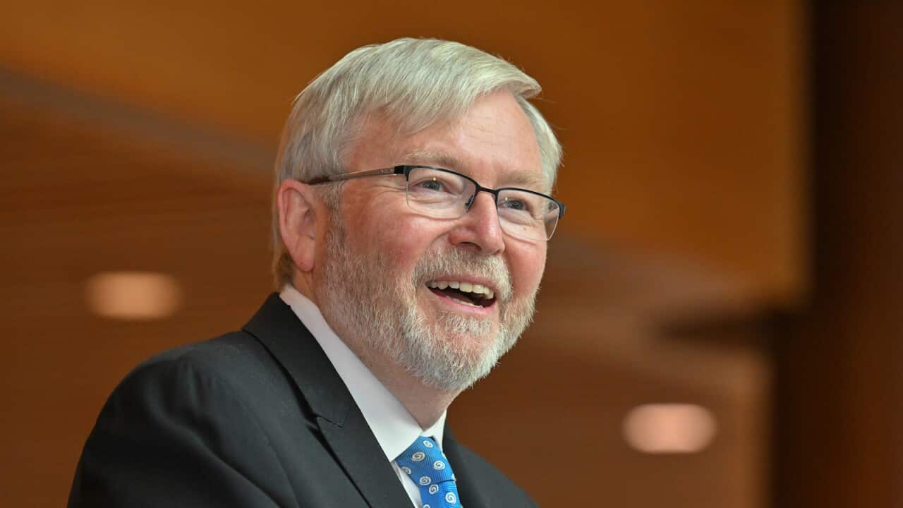 Kevin Rudd wearing a suit and smiling in front of a blurred brown wall.