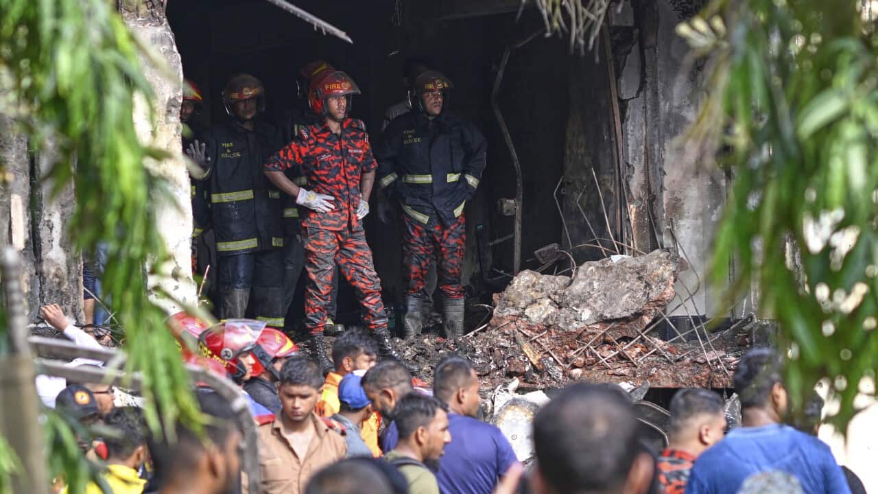 Firefighters stand in rubble of building surrounded by crowds of people.