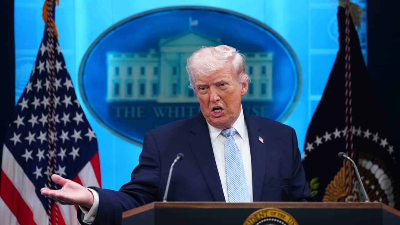 A man wearing a suit stands behind lectern with arm outstretched