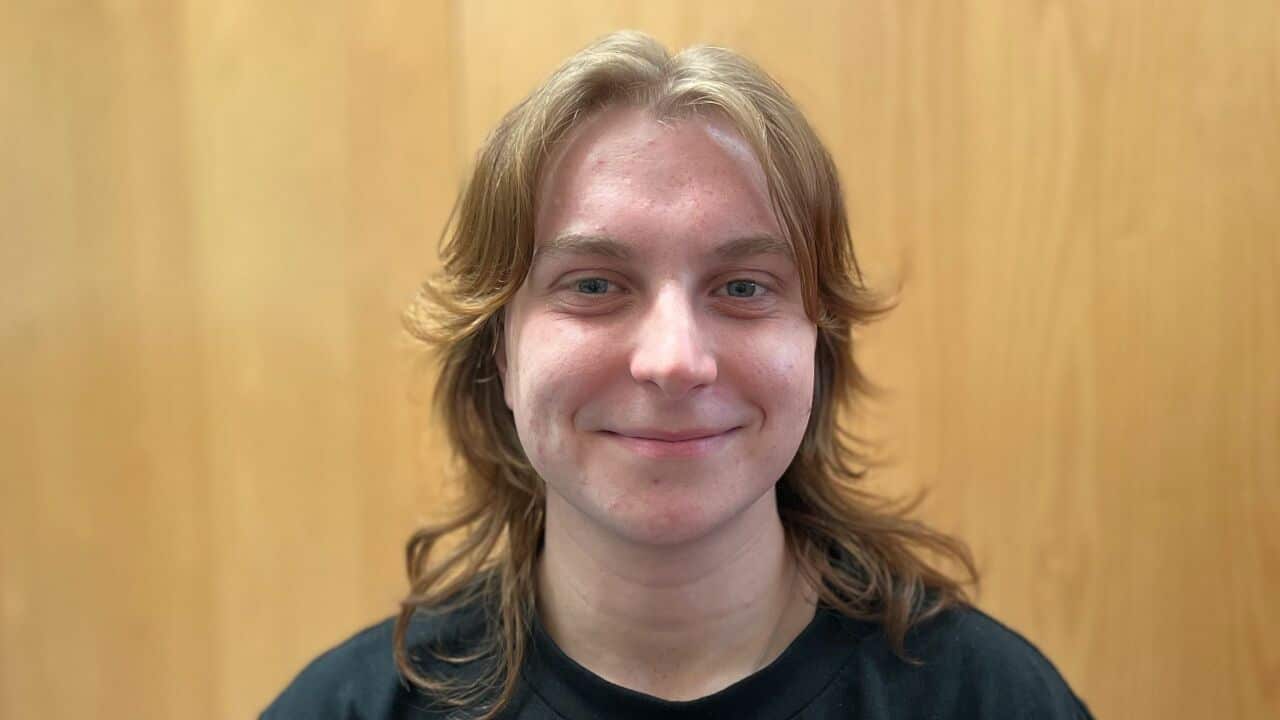 a close up shot of a young man with a shaggy blonde haircut in a black t shirt in front of a timber background