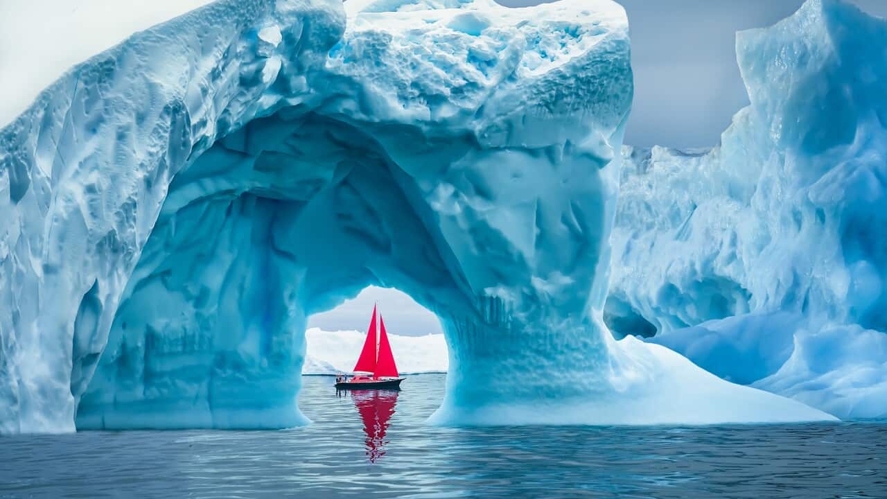 Red sailboat sailing under an iceberg arch on sunny blue Arctic Ocean in Greenland (Getty).
