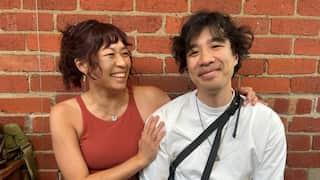 A man smiles at the camera with a woman looking at him and laughing and holding him