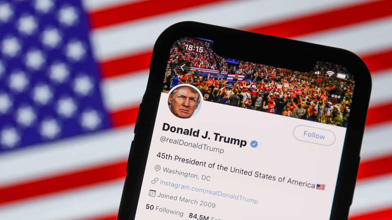 Donald Trump's Twitter profile on a mobile phone screen in front of an American flag.