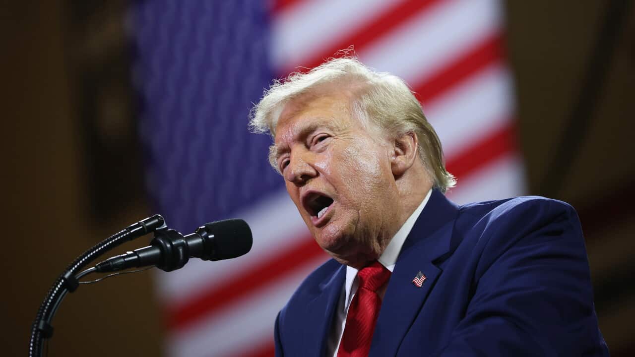 Donald Trump in a navy suit and red tie speaking into a microphone.