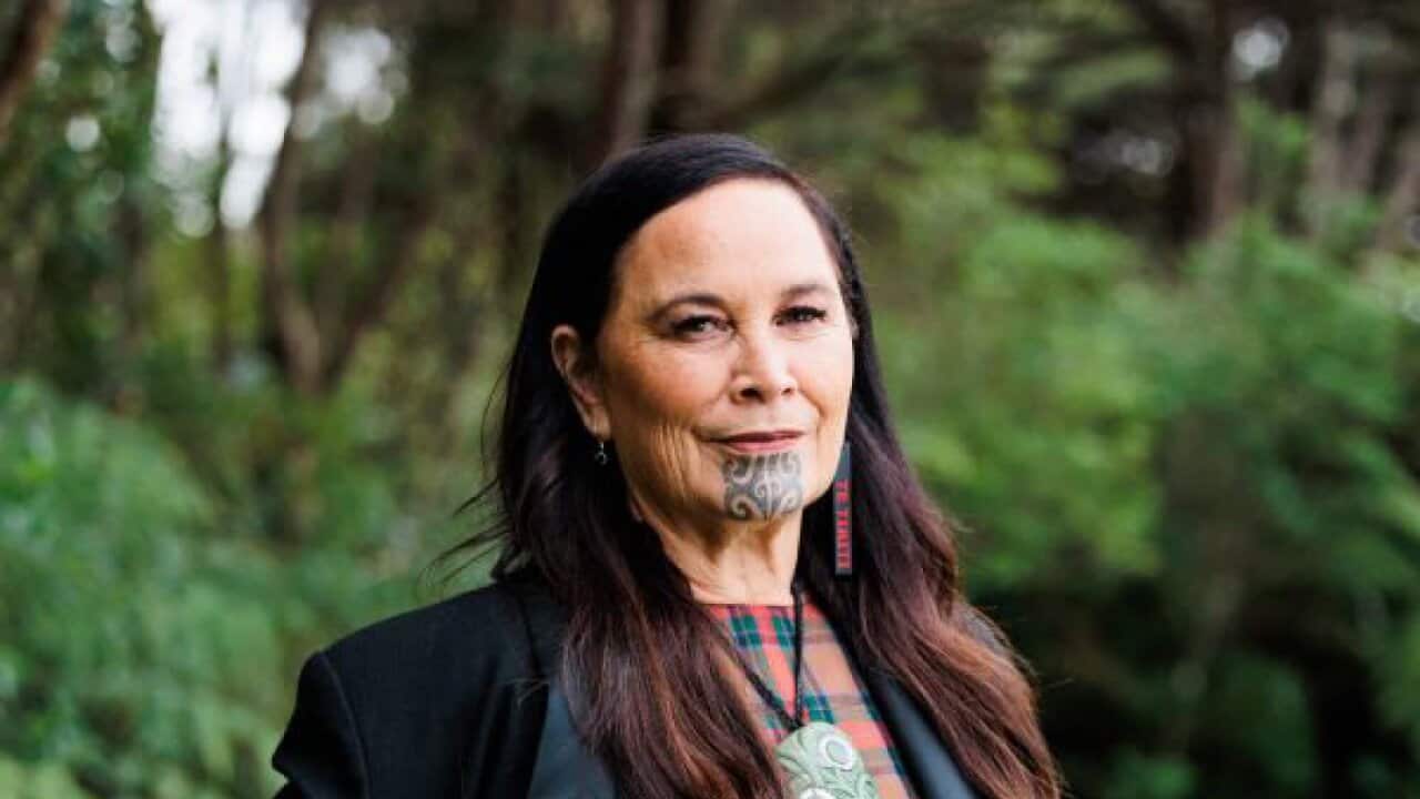 A middle-aged woman with long brown hair and a Māori Moko Kauae (facial tattoo) stands in front of some green trees.