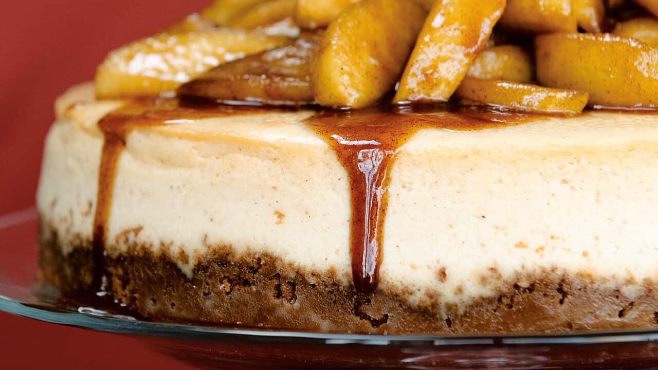Part of the side of a creamy white cheesecake is seen close-up. It has a brown base and is topped with pieces of apple in a sticky sauce. A little of the sauce has run down the side.