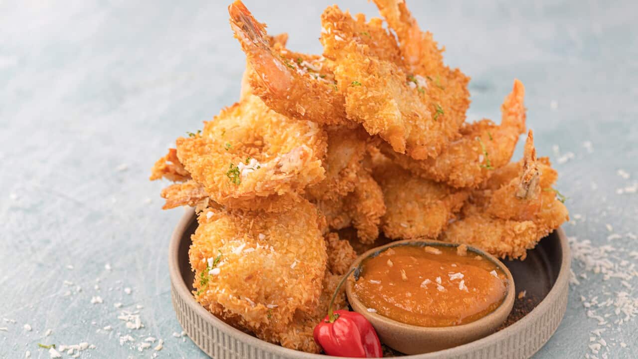 A stack of golden crumbed prawns sits in a round tray with a shallow side, along with a small chilli and a small bowl of orange-ish sauce.