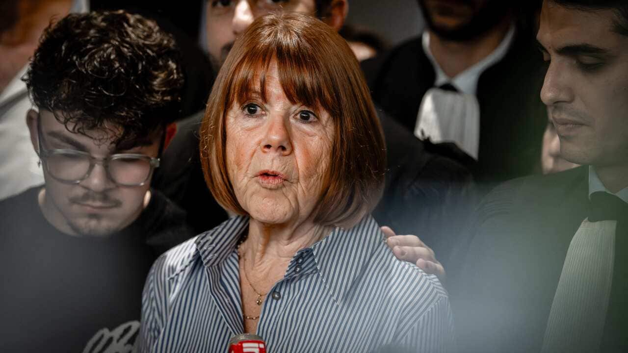 A woman in a striped shirt stands next to younger men speaking into a microphone.