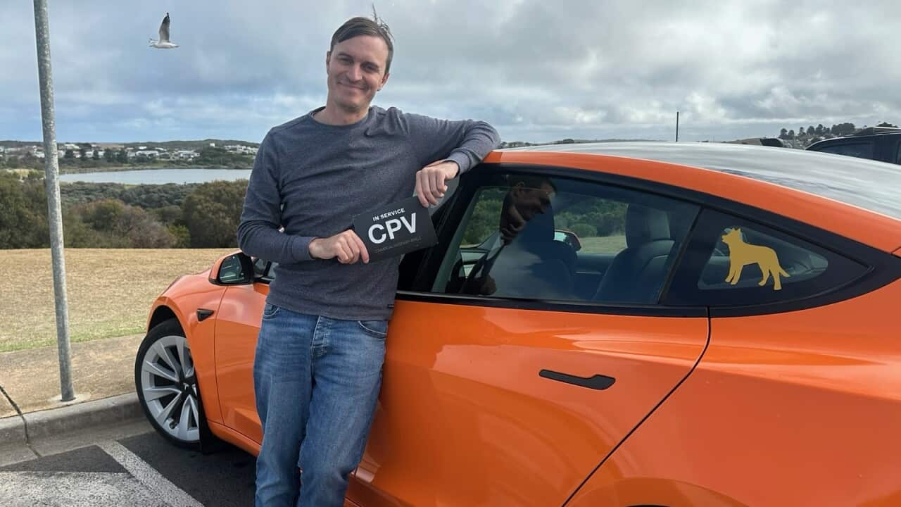 A man wearing a grey jumper and blue jeans leans against a bright orange car.