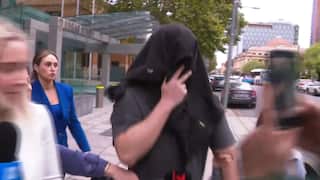 A person with a black cloth draped over their head and face walks past a crowd of journalists and cameras outside a building.