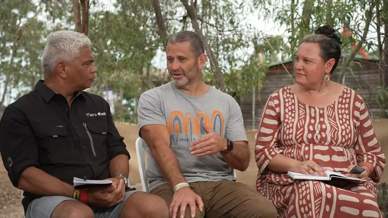 Three people seated outdoors in a panel setting, including an NITV reporter, engaged in an earnest conversation.