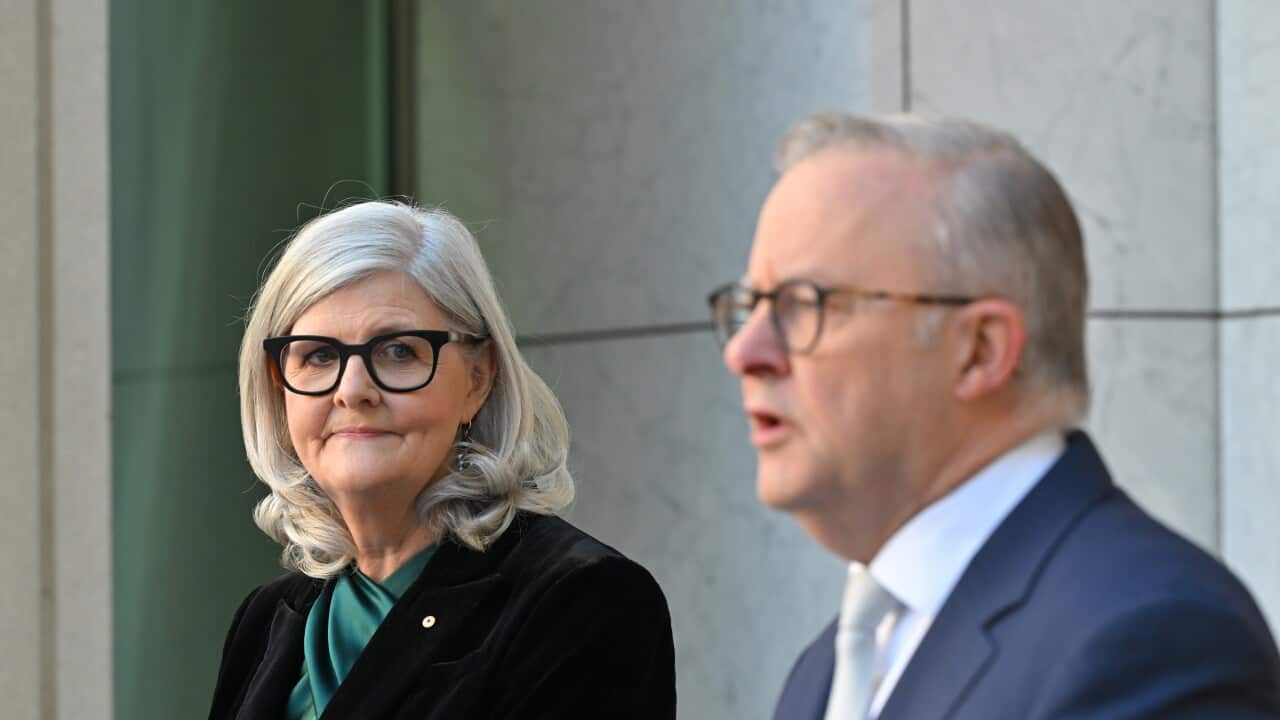 A woman with grey hair and glasses looks to her left at a man in a suit speaking at a lectern.