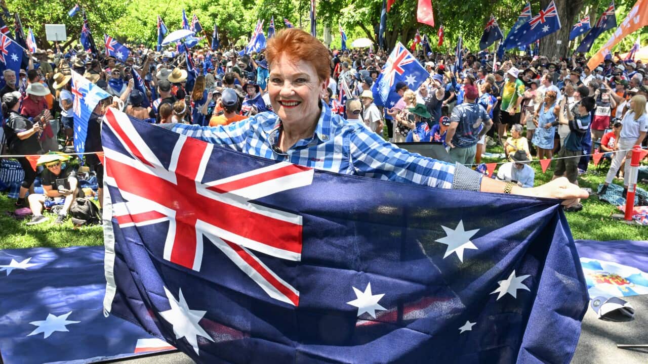 Australian politician Pauline Hanson smiles while holding up a large Australian flag in front of a crowd of supporters at an outdoor rally.