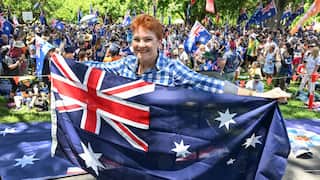 Australian politician Pauline Hanson smiles while holding up a large Australian flag in front of a crowd of supporters at an outdoor rally.