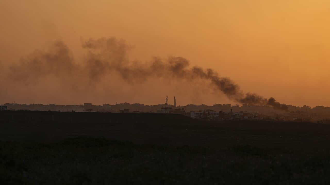 Smoke billows over the horizon, where buildings can be seen in the distance with an orange sky.