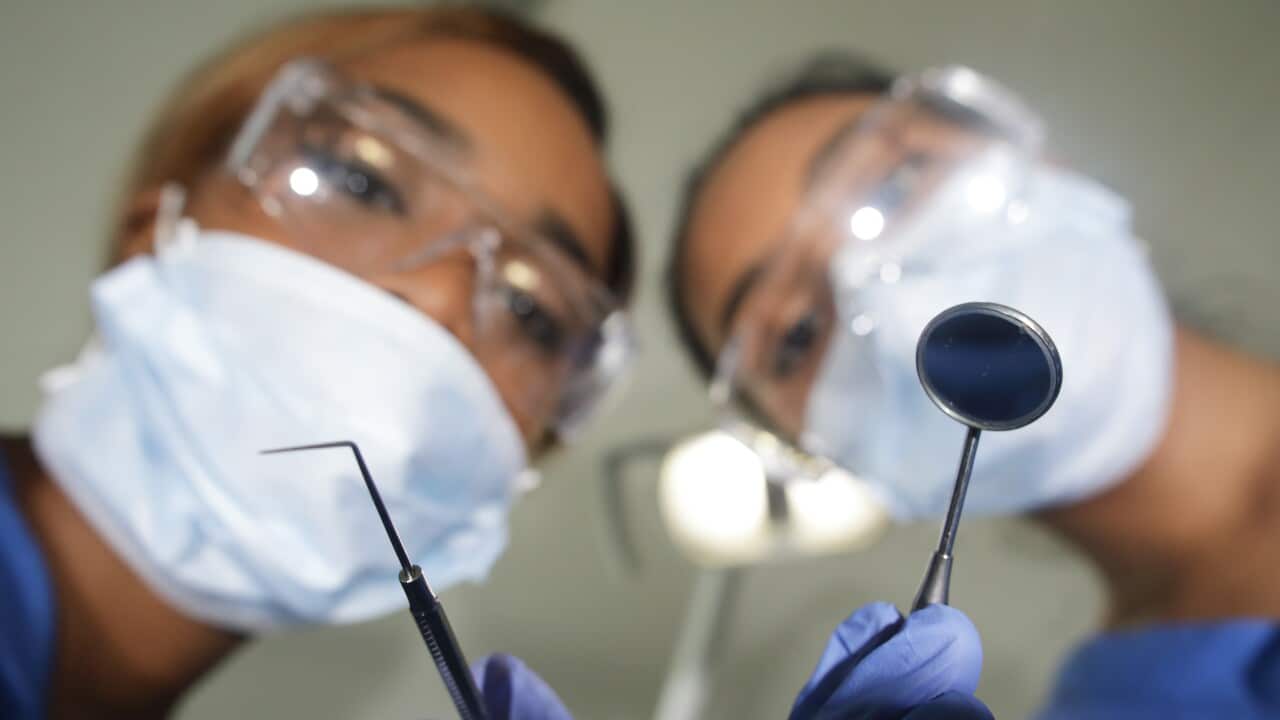Two female dentists with dental instruments in their hands, looking down. They are wearing glasses and masks.