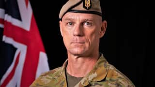 A middle-aged white man in military uniform stares ahead in front of an Australian flag.