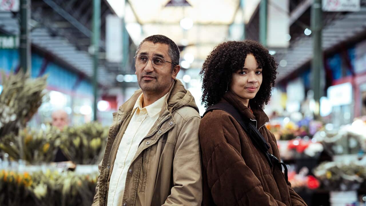 A man and a woman stand back to back in what looks like a flower market.