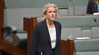 A blonde woman in a striped grey suit with a white shirt walks past a row of plush green bench seats.