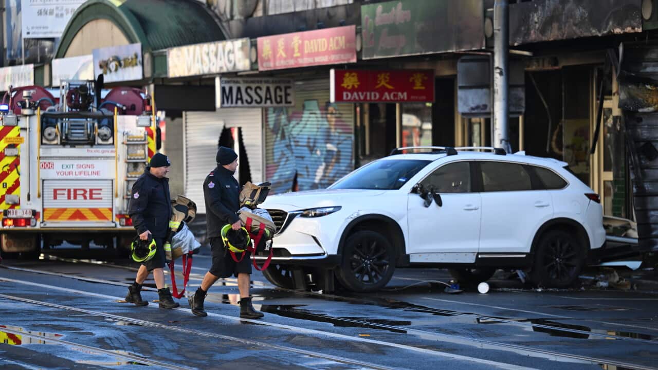 Two firefighters carry gear across a wet city street past a fire engine and a white SUV parked in front of fire-damaged storefronts.