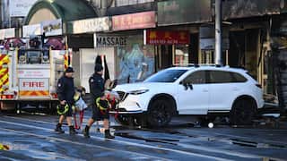 Two firefighters carry gear across a wet city street past a fire engine and a white SUV parked in front of fire-damaged storefronts.