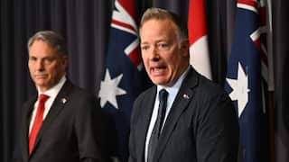 Two middle aged men in suits stand before Australian flags and a black curtain. One of the two men is speaking.