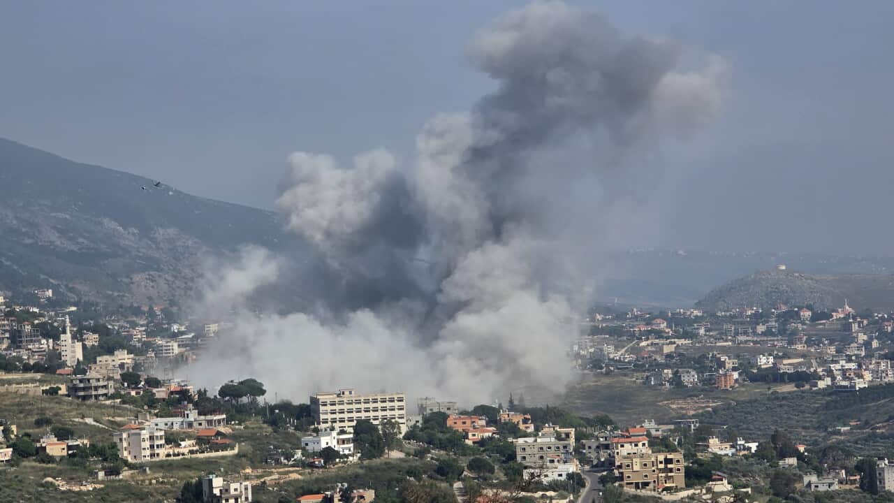 Large plumes of smoke rise above a residential area following an apparent airstrike, with buildings and surrounding hills visible in the background.