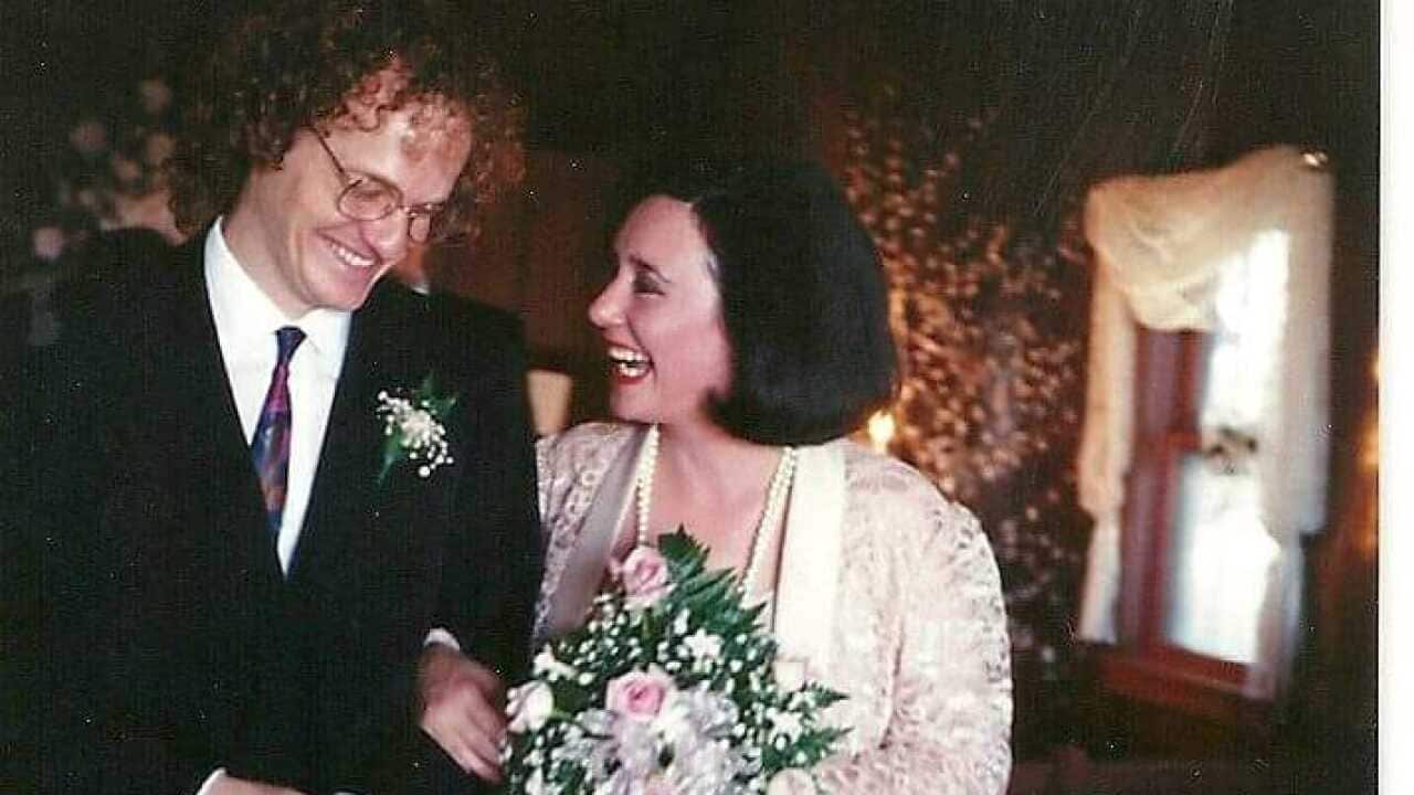 Colette holds a bouquet of flowers, linking arms with her husband at their wedding.