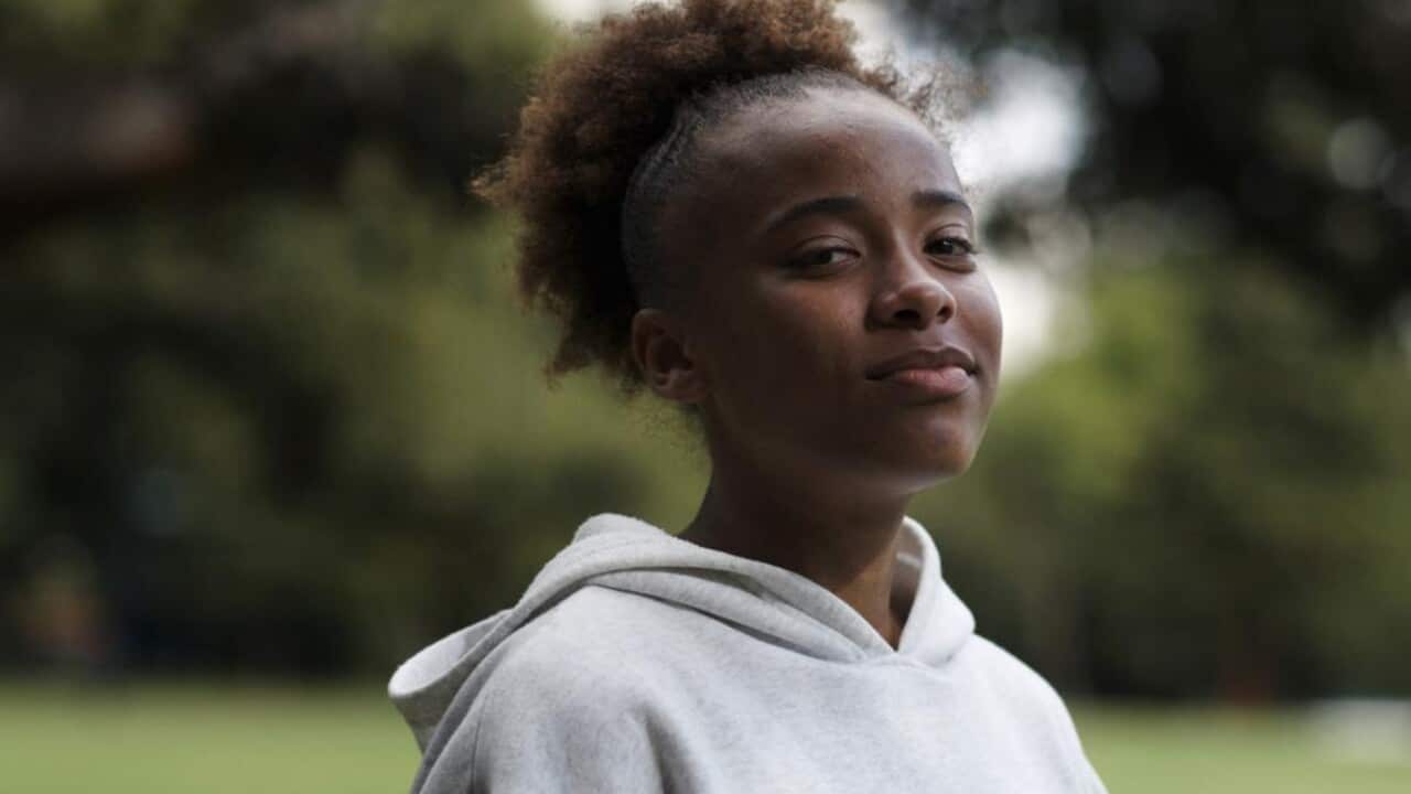 A young woman in a light grey jumper looks at the camera while at the park.