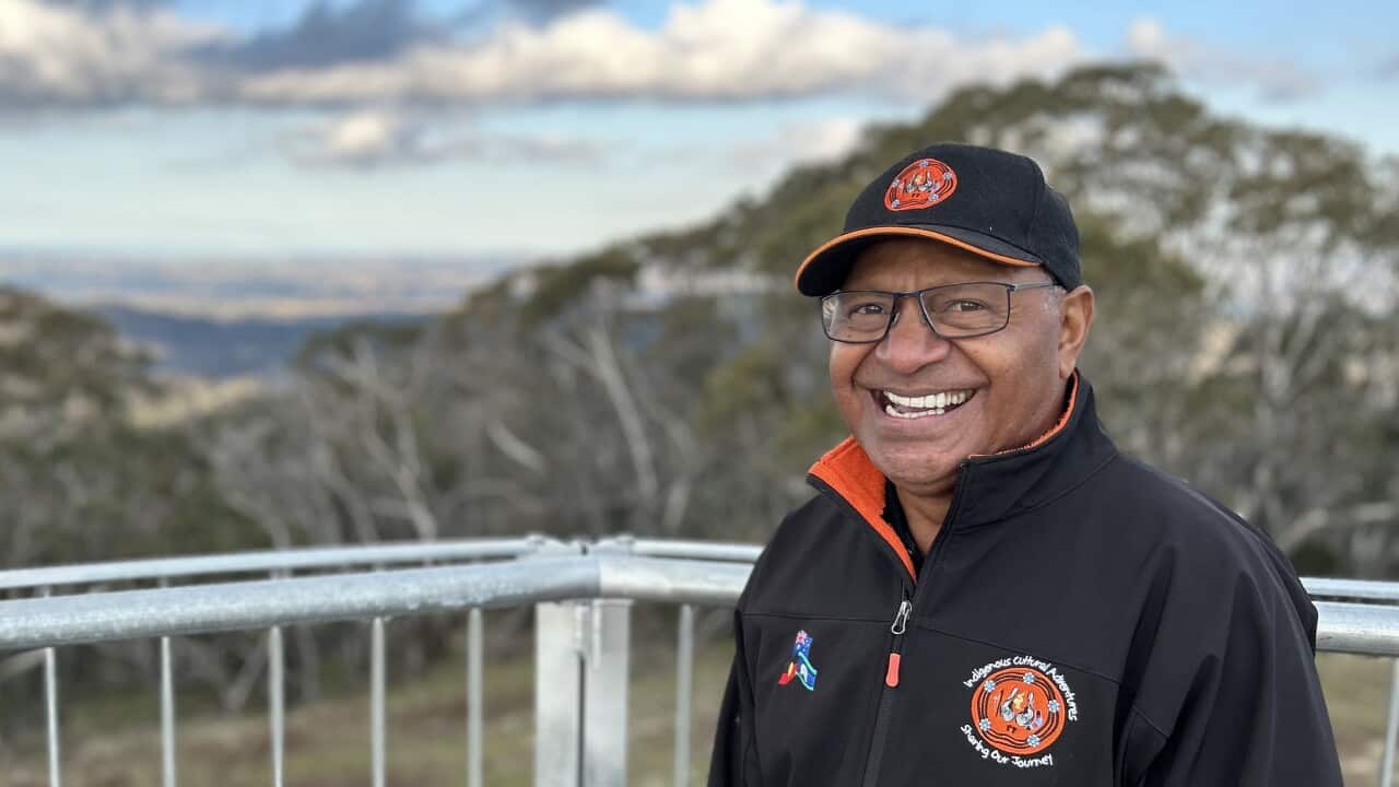 A man wearing a dark jacket and cap and glasses smiles while standing overlooking a rural valley.
