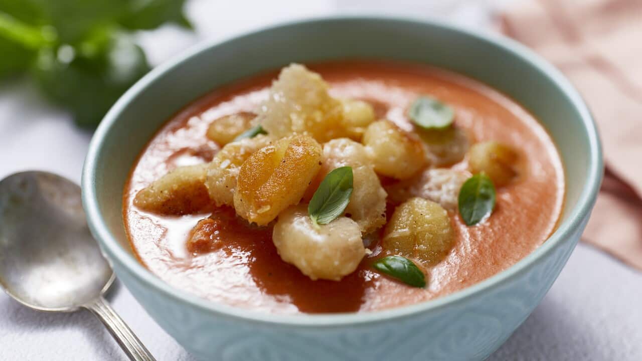 A blue soup bowl with a patterned outside surface holds tomato soup topped with a tumble of cooked gnocchi and small basil leaves. A burnished silver soup spoon sits alongside.