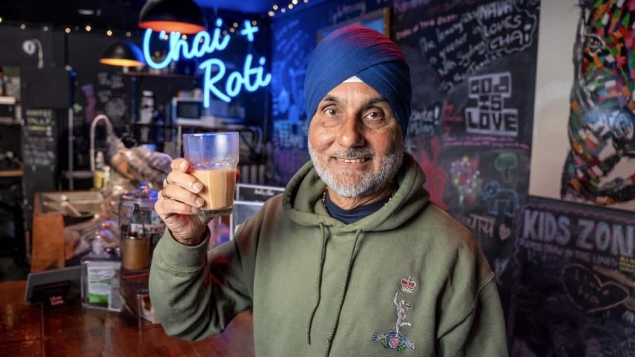 a man wearig a blue turban and a green sweater stands in a bar holding a cup of tea.