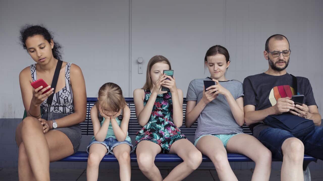 Adults and children sitting on a bench distracted by mobile phones whilst younger child without a phone is completely fed up