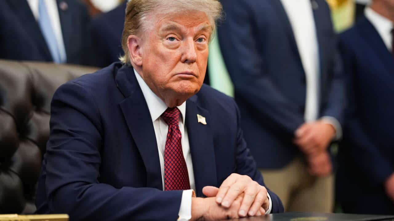 US President Donald Trump sits at a desk in the White House with his hands clasped as officials stand behind him during a meeting.