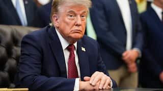 US President Donald Trump sits at a desk in the White House with his hands clasped as officials stand behind him during a meeting.