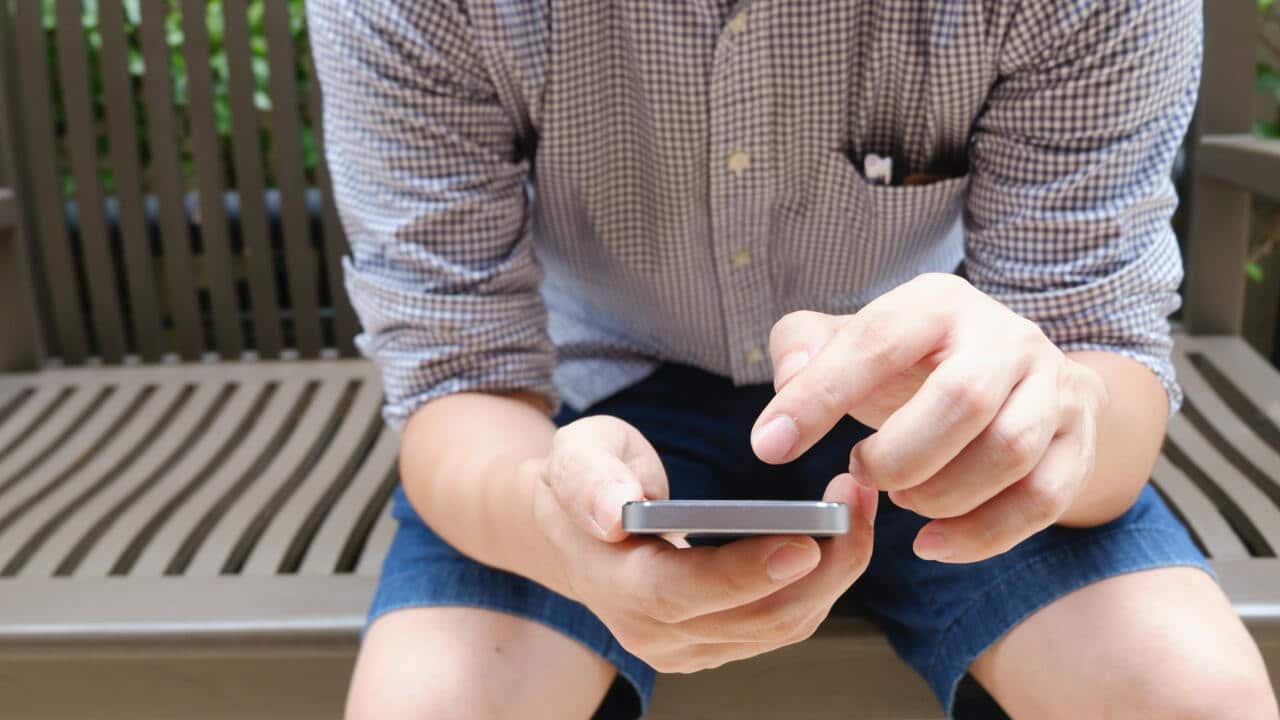 Man Wearing Brown and White Plaid Sport Shirt Sittings on Brown Bench and Using Smartphone during Day