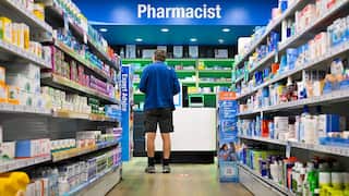 A man in a blue shirt and black shorts stands at a pharmacist's counter.